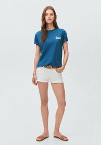 Modré bavlněné tričko s kulatým výstřihem a krátkými rukávy, spárované s bílými džínovými šortkami. Doplněno hnědými pantoflemi a páskem.