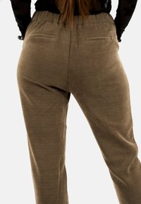 Persona con pantaloni di velluto a coste marroni vista di spalle, con le mani sui fianchi e i lunghi capelli che cadono su una maglietta nera.