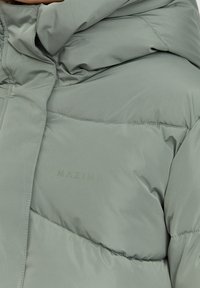 Pufferjakke i lys grøn, med en glat, mat tekstur og vandret syninger. Brandlogoet "MAZINE" er præget på forsiden.