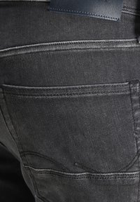 Tela de denim gris oscuro con detalles de bolsillo cosidos. Presenta una etiqueta de cuero y una textura ligeramente desgastada. Vista en primer plano.