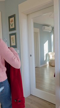 Červený vlněný kabát zavěšený na ramínku, světle růžový svetr, dřevěné podlahy, dva zarámované botanické tisky na světle modré stěně, velké zrcadlové dveře.