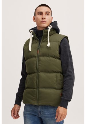 Vestă puffer de culoare verde măslin, cu fermoar în față, două buzunare laterale și glugă. Pulover texturat negru purtat dedesubt, cu detalii de tricot vizibile.