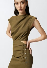Haljina maslinasto zelene boje s kratkim rukavima, s nabranom stranom, prorezom i ukrasnim metalnim gumbima uz bočni šav.
