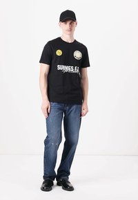 T-shirt en coton noir avec le texte "SUNNIES E.C." en blanc et un logo circulaire. Porté avec un jean en denim bleu, des chaussures noires et une casquette noire.