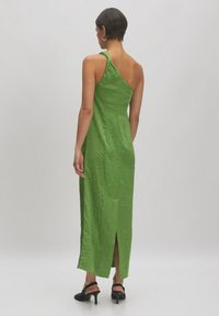 Grünes, strukturiertes, ärmelloses Kleid mit drapiertem Schulterbereich und seitlichem Schlitz. Gestylt mit schwarzen, hohen Sandalen, von hinten gezeigt.