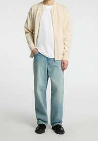 Persona vestida con una chaqueta cremosa con cremallera, camiseta blanca, pantalones vaqueros sueltos de color azul claro y zapatos negros, de pie con una mano en el bolsillo.