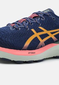 Gros plan sur une chaussure de course bleu marine avec un logo orange, une semelle rose et grise, et des lacets bleu marine avec des accents orange.