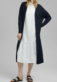 Marineblauer langer Cardigan über einem weißen, gestuften Kleid. Der Cardigan hat einen lässigen Schnitt, eine offene Front und lange Ärmel, kombiniert mit schwarzen Sandalen.