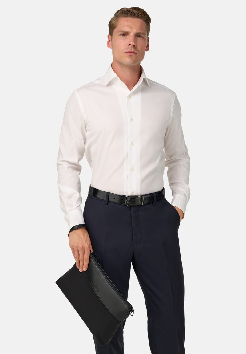 Camicia bianca con maniche lunghe, abbinata a pantaloni eleganti blu navy. Clutch nera in pelle tenuta in una mano, cintura nera che mette in risalto la vita.