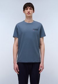 Camiseta de algodón en azul pizarra con mangas cortas, cuello redondo y un pequeño logotipo en negro y rojo. Textura suave, corte relajado.