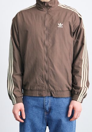 Persoon draagt een bruin Adidas jack met rits en beige strepen op de mouwen en een blauwe spijkerbroek, staand tegen een effen achtergrond.