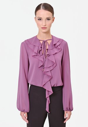 Donna che indossa una camicetta a maniche lunghe color malva con volant sovrapposti e un fiocco legato sulla scollatura, abbinata a pantaloni neri.