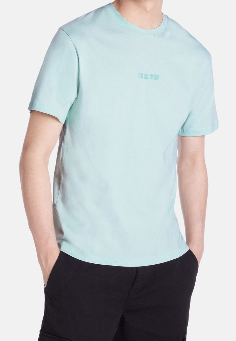 Personne portant un t-shirt bleu clair à manches courtes avec le texte "THE KOOPLES" sur la poitrine et un pantalon noir, les mains dans les poches, sur fond blanc.