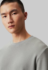 Suéter gris texturizado con cuello redondo, que presenta un suave patrón de punto acanalado. Primer plano de la cara y la parte superior del cuerpo del modelo.