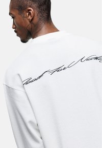 White sweatshirt with a textured finish, featuring black cursive embroidery on the back reading "Rest For Nothing."