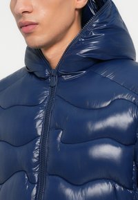 Jachetă puffer bleumarin cu un finisaj lucios, având un fermoar frontal, o glugă și modele ondulate quilted pe material.