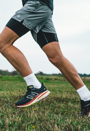 Des chaussures de sport noires avec des semelles blanches et des accents verts, associées à un short gris et des chaussettes blanches, portées par une personne courant sur l'herbe.