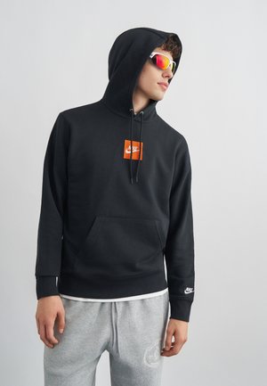 Fiatal férfi fekete Nike kapucnis pulóverben, szürke melegítőnadrágban és tükröződő sport napszemüvegben, oldalra néz egy egyszerű világos háttér előtt.