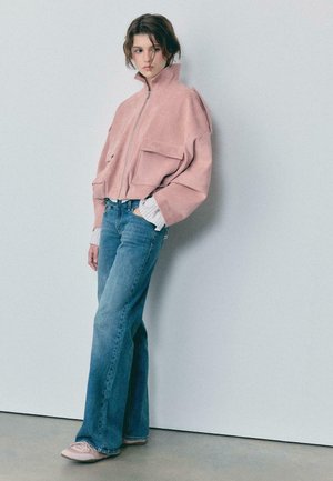 Donna che indossa una giacca rosa oversize con cerniera, jeans larghi blu e sneakers rosa, in piedi contro un muro grigio chiaro uniforme.