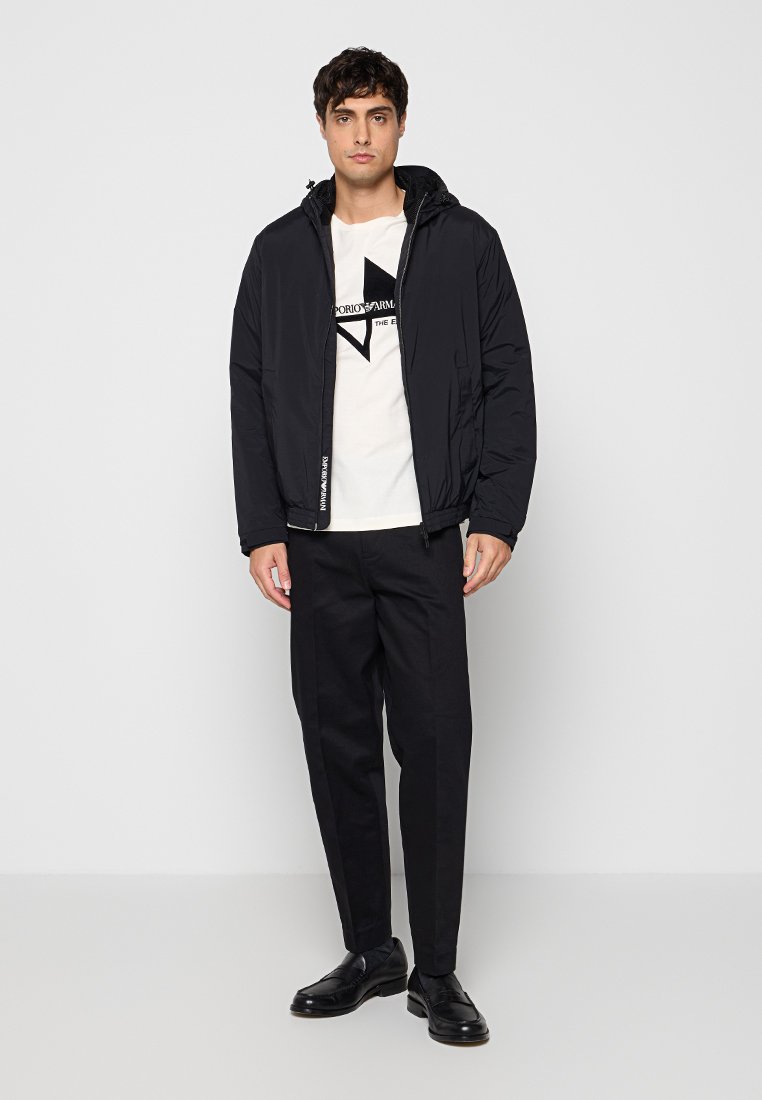 Joven con chaqueta negra con capucha, camiseta blanca con diseño gráfico, pantalones negros y zapatos de vestir negros, de pie frente a un fondo claro y liso.