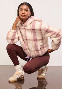 Bolyhos kockás kapucnis pulóver bézs és rózsaszín árnyalatokban, elülső zsebbel, húzózsinórral a deréknál és puha textúrával, barna leggings-szel és sportcipővel párosítva.