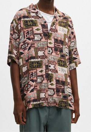 Mężczyzna ubrany w luźną, krótką koszulę na guziki z różowymi, czarnymi i zielonymi abstrakcyjnymi geometrycznymi wzorami oraz ciemnozielone spodnie.