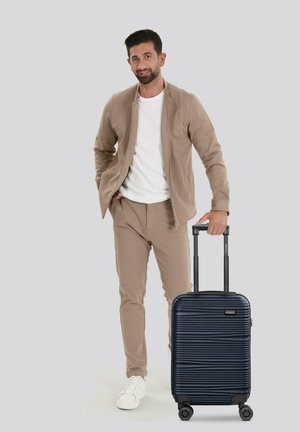 Blauer Hartschalenkoffer mit horizontalen Rillen, ausziehbarem Griff und vier Rädern. Ein Modell in beiger Kleidung steht daneben.