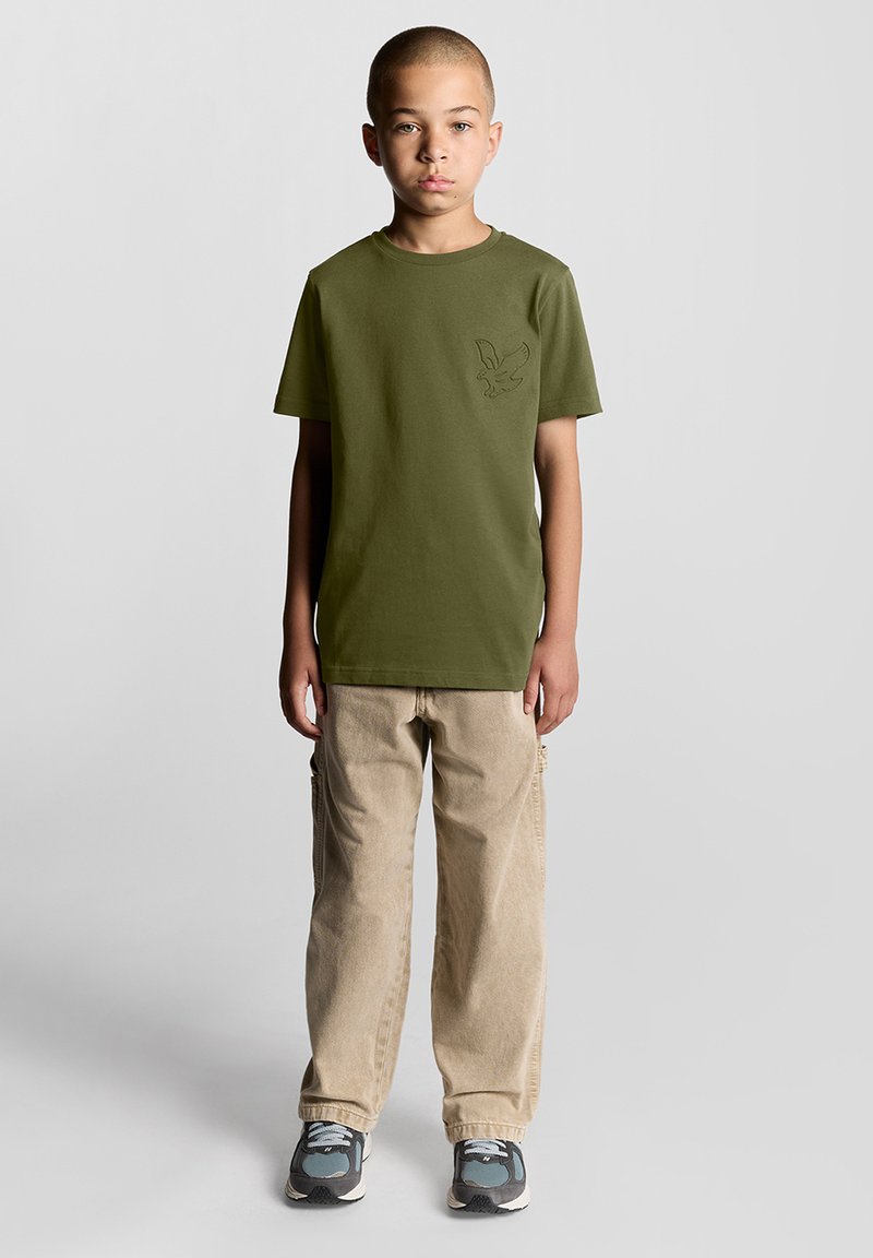 Olivengrøn bomulds T-shirt med korte ærmer, prydet med et diskret ørne-grafik. Kombineret med beige cargo-bukser og sneakers.