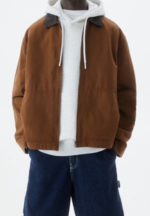 Veste en coton marron avec un col foncé, deux poches avant, superposée à un hoodie gris avec des cordons, associée à un short bleu foncé.