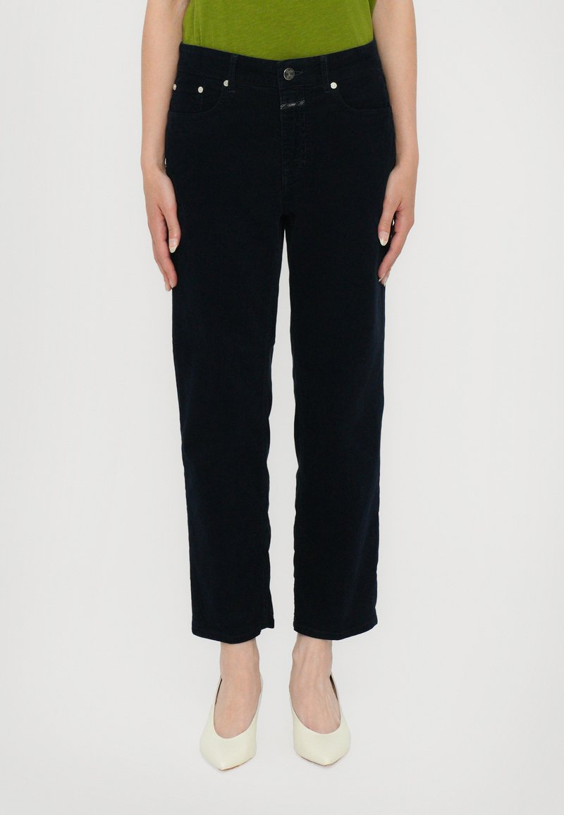 Navy blue jeans with a straight leg design, featuring five pockets, front button closure, and a smooth texture. Paired with white high heels.