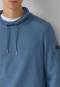 Niebieski sweter z wysokim kołnierzem, prążkowanymi detalami na ramionach i sznureczkami do ściągania. Zawiera naszywkę z logo na rękawie. Wykonany z bawełnianej mieszanki.