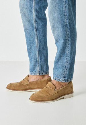 Mocassini in pelle scamosciata color marrone chiaro con design slip-on, caratterizzati da una sottile lavorazione a punti, punta arrotondata e suola in gomma bianca, abbinati a jeans celesti.