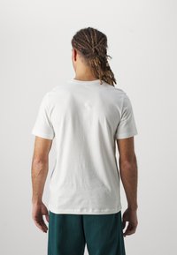 Camiseta blanca de algodón con mangas cortas, diseño de cuello redondo, textura suave, costuras visibles, combinada con pantalones verdes, vista desde la parte posterior.