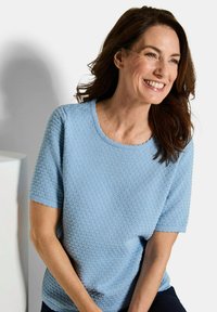 Lachende vrouw met lang bruin haar, gekleed in een gemêleerd lichtblauw t-shirt met korte mouwen, zittend tegen een eenvoudige witte achtergrond.