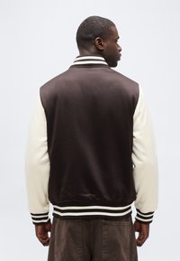 Άνδρας με την πλάτη γυρισμένη φοράει ένα καφέ varsity μπουφάν με λευκά μανίκια και ριγεμένη ριμπέ ζώνη, γιακά και μανσέτες, πάνω σε απλό φόντο.