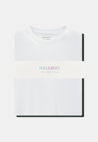 White cotton t-shirt with a round neck, folded with a black shirt behind it. Features a text band reading "PULL&BEAR" and slogan.
