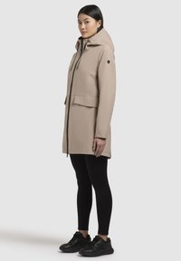 Beige hættefrakke med frontlynlås, store lommer og sorte detaljer, parret med sorte leggings og sneakers. Glat tekstur.