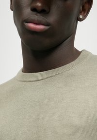 Maglione a maglia testurizzata verde chiaro con scollatura rotonda, caratterizzato da un colletto a costine e una vista ravvicinata del tessuto e del tono della pelle.