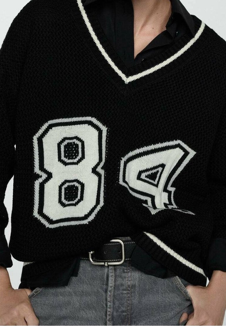 Pull noir tricoté oversize avec des accents blancs présentant les chiffres 84 et 4. Design à col en V, tissu texturé, porté sur une chemise noire.