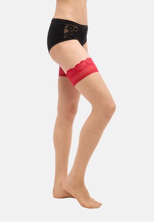 Kvinde iført sorte trusser med blonder og røde lårhøje blonderstrømper, stående på tæer mod en hvid baggrund.