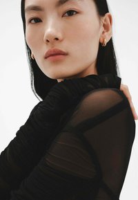 Top negro transparente con mangas largas fruncidas, que presenta una textura sutil. La modelo tiene una piel lisa y lleva pequeños pendientes de aro dorados.