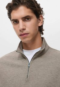 Giovane uomo con capelli ricci scuri che indossa un maglione beige con cerniera sopra una camicia bianca, che guarda direttamente nella fotocamera con un'espressione neutra.