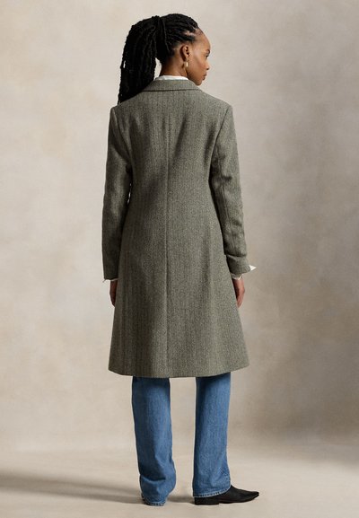 Femme aux cheveux tressés portant un long manteau gris en chevrons, un jean bleu et des chaussures noires, debout de dos sur un fond neutre.
