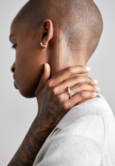 Silver hoop earring and a delicate silver ring on a hand, with skin showing lace-like tattoos. Soft gray sweater texture visible.