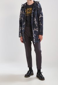 Bunda s maskáčovým vzorem v námořnické modři a šedé barvě, s kapucí a zapínáním na zip, ve spojení s tmavými kalhotami a černými šněrovacími botami.