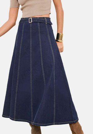 Spódnica jeansowa w kształcie litery A o wysokim stanie w ciemnoniebieskim kolorze, z pionowym przeszyciem, dopasowanym paskiem z kwadratową klamrą, noszona z beżowymi botkami i złotą bransoletką na nadgarstek.