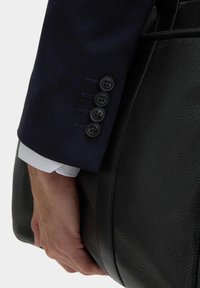 Czarna skórzana torebka trzymana w dłoni, z teksturowanym wykończeniem i gładkim skórzanym paskiem. Częściowo widoczna marynarka w granatowym kolorze z guzikami.