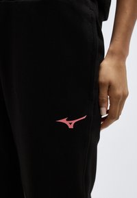 Κοντινό πλάνο ενός χεριού που ακουμπά δίπλα σε μαύρο παντελόνι με μικρό ροζ λογότυπο Mizuno στον μηρό.