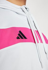 Weißer Hoodie mit einem pinken horizontalen Streifen und einem schwarzen Adidas-Logo. Glatte Textur mit verstellbaren Kordeln am Hals.
