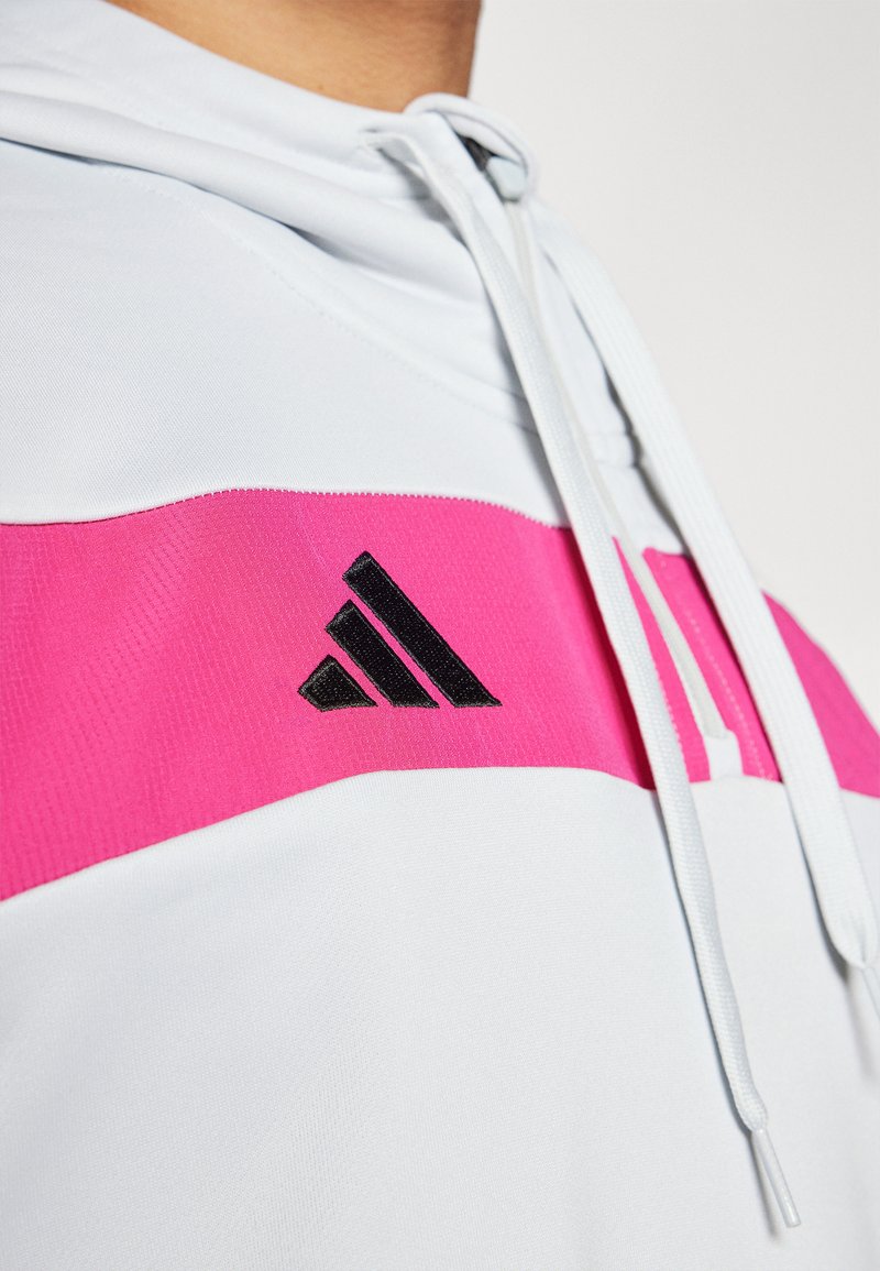 Sudadera blanca con una franja horizontal rosa que presenta un logo de Adidas en negro. Textura suave con cordones ajustables en el cuello.